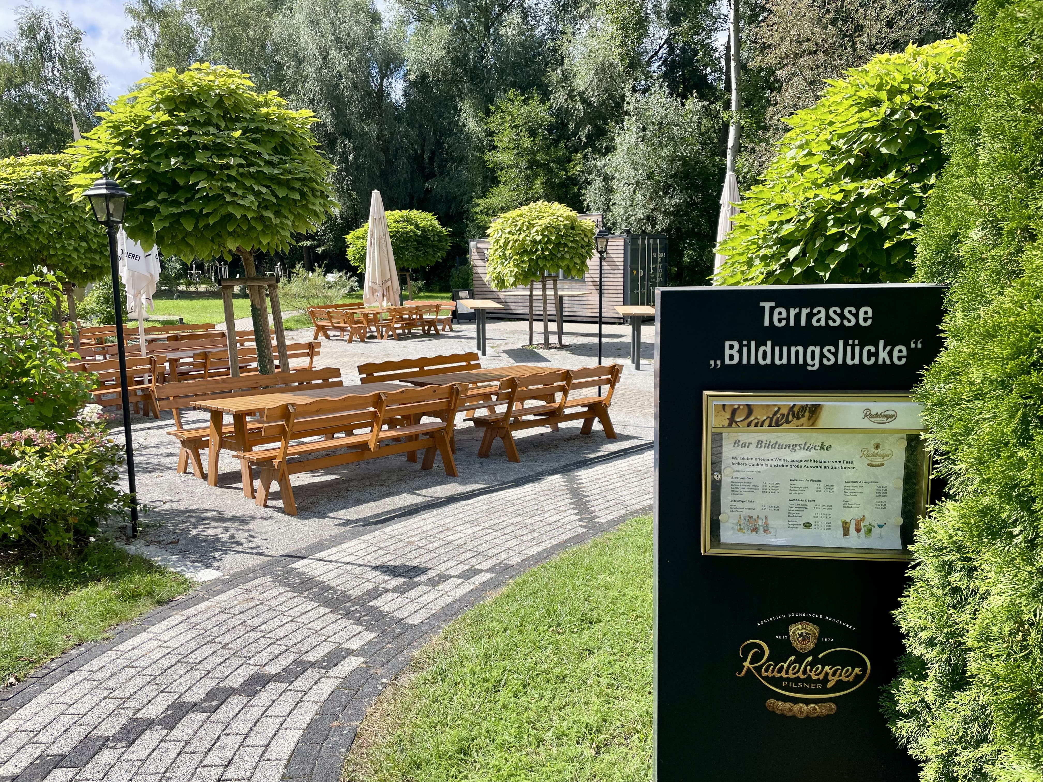 Sonnige Terrasse mit Holzbänken zwischen Bäumen, davor eine Tafel mit der Aufschrift „Terrasse Bildungslücke“.