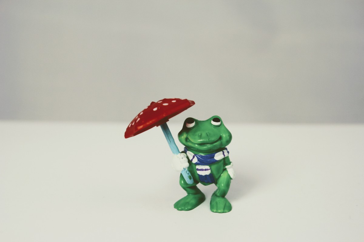 Grüne Froschfigur mit blau-weiß gestreiftem Schal, die einen roten, weiß gepunkteten Schirm hält