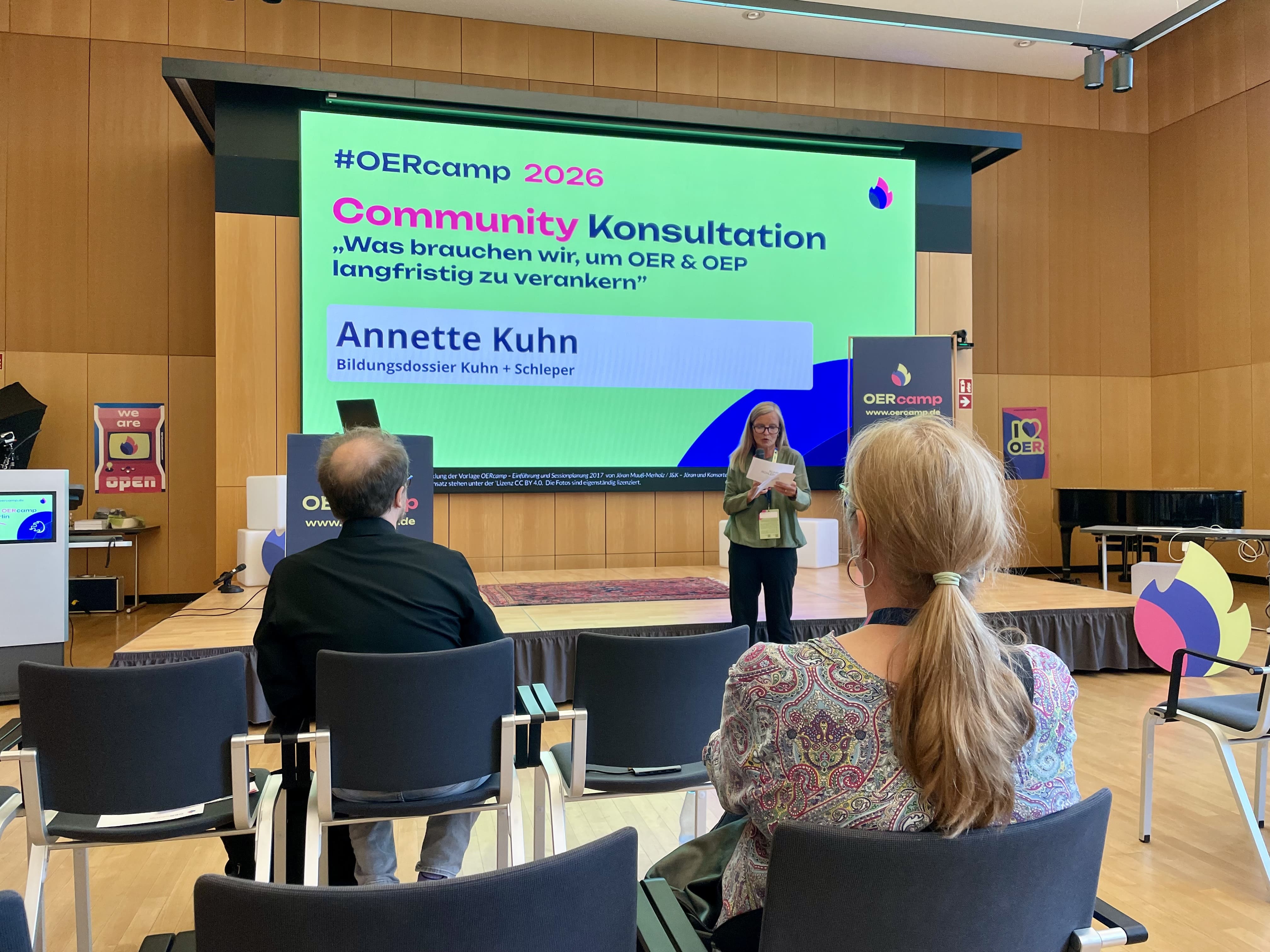 Blick aus dem Publikum auf die Bühne: Annette Kuhn eröffnet die Community-Konsultation, auf der Leinwand die Frage „Was brauchen wir, um OER & OEP langfristig zu verankern“.