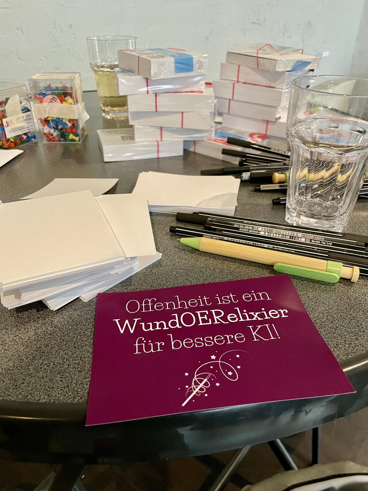 Stehtisch mit verschiedenen Materialien wie Stifte, Zettel und Stecknadeln. Darauf liegend Postkarte mit Abbildung von Zauberstab und Aufschrift “Offenheit ist ein WundOERelexier für bessere KI”.
