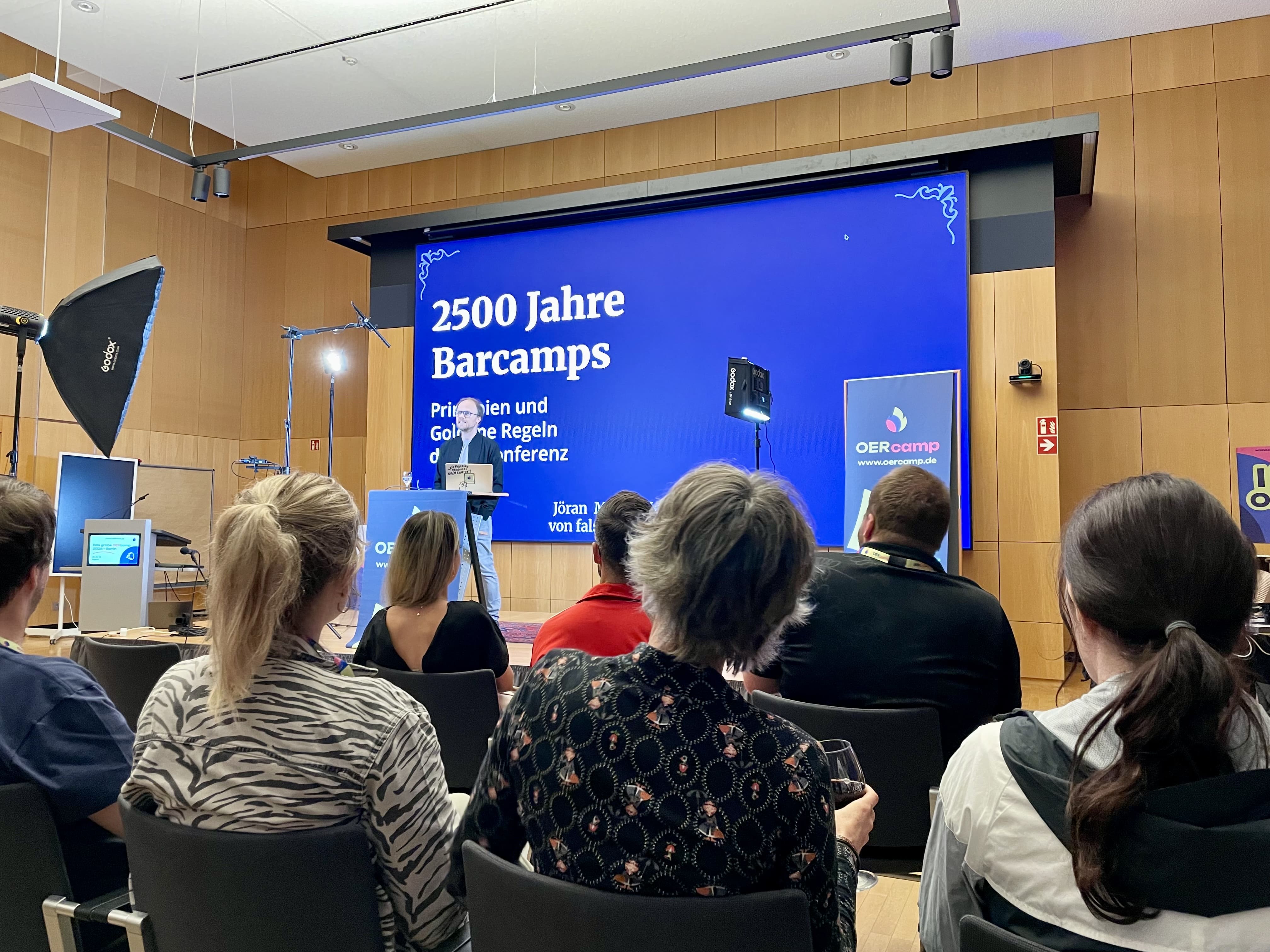 Jöran Muuß-Merholz am Rednerpult vor einer Folie mit dem Titel „2500 Jahre Barcamps“, im Vordergrund Zuhörende von hinten.